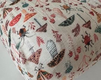 May include: A cream-colored fabric with a repeating pattern of insects, mushrooms, and foliage in shades of red, orange, green, and brown. The fabric has a textured appearance and a soft, natural aesthetic.