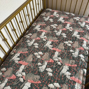 May include: A crib sheet with a dark green background featuring a repeating pattern of brown frogs sitting on white and red mushrooms. The mushrooms have white stems and red caps with white spots.