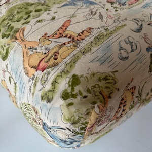 Puede incluir: Una tela estampada con un diseño repetitivo de Winnie the Pooh y Tigger en una escena al aire libre. El diseño incluye ilustraciones de los personajes, árboles y otros elementos en colores pastel suaves.