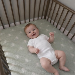 May include: A baby in a white onesie smiles while lying in a crib. The crib has a metal frame and a light green patterned sheet. The baby has red hair.