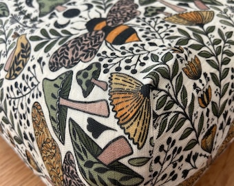 May include: Close-up of a fabric with a whimsical pattern featuring bees, mushrooms, and foliage in shades of green, brown, and orange on a cream background. The design is detailed and nature-inspired, suitable for home decor or craft projects.
