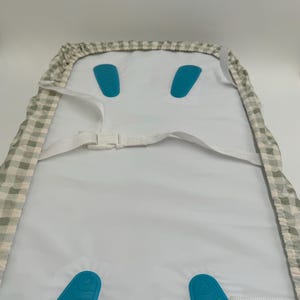 May include: A white high chair cover with a grey and white checked pattern. The cover has two blue straps with buckles and two blue padded inserts.