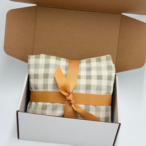 May include: A white gift box with a brown cardboard lid. The box is open and contains a green and white gingham fabric wrapped with a gold ribbon.