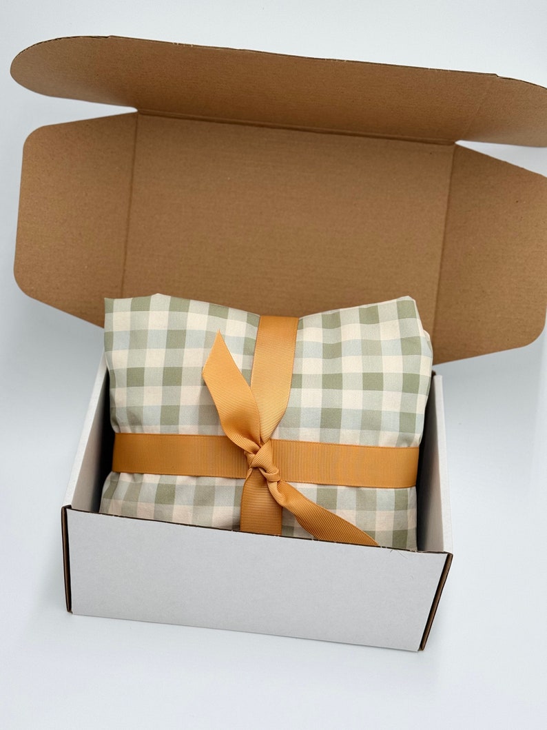 May include: A white gift box with a brown cardboard lid. The box is open and contains a green and white gingham fabric wrapped with a gold ribbon.