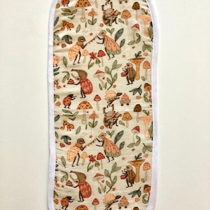 Peut inclure: Une manique de four ovale blanche avec une bordure blanche. La manique est recouverte d'un tissu beige avec un motif de bêtes brunes, orange et rouges, de champignons et de feuilles.