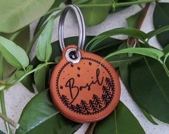 May include: A brown leather keychain with a circle-shaped tag. The tag has a black and white design with a stylized mountain range and the word "Basil" written in cursive.