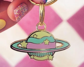 May include: Gold-toned keychain featuring a colorful planet charm. The planet has a light green, pink, and teal design with star accents. The background is a pink and white checkered pattern.