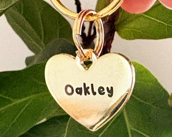 May include: A gold-toned heart-shaped metal tag attached to a circular split ring. The front of the heart features the name Oakley engraved in a dark, clear font. The metal surface has a polished, reflective finish, catching light across its smooth, curved edges.
