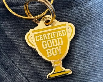 May include: A gold-colored keychain with a trophy-shaped charm. The charm has the words "CERTIFIED GOOD BOY" in yellow lettering. The keychain is attached to a gold ring.