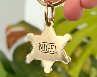 May include: A gold-toned metal keychain in the shape of a six-pointed sheriff star badge. The center of the badge is engraved with the name NIGEL in capital letters, flanked by two horizontal lines above and below the text. It has a circular ring.
