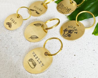 May include: Six gold-toned keychains with different designs and names. The keychains feature a mushroom design with the name "Mushy", a leaf design, a hot dog design with the name "Wesley", a butterfly design with the name "Oslo", a landscape design, and a leaf design with the name "Bubba".