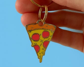 May include: A pizza slice keychain with a gold-colored ring and clasp. The pizza slice has a yellow base with red pepperoni and orange crust. The keychain is held against a light blue background.