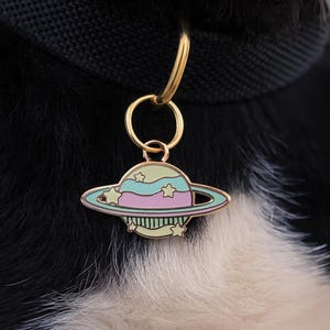 May include: A colorful planet-shaped pet tag with a gold-colored ring. The tag features a light blue, pink, and green planet design with star accents. The tag is attached to a black pet collar, set against a background of black and white fur.
