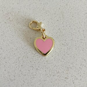 May include: A gold-colored heart charm with a pink enamel center. The heart-shaped pendant is attached to a gold-colored lobster clasp. The charm is a jewelry accessory.