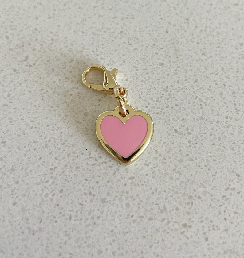 May include: A gold-colored heart charm with a pink enamel center. The heart-shaped pendant is attached to a gold-colored lobster clasp. The charm is a jewelry accessory.