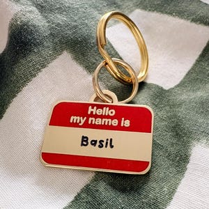 May include: A rectangular gold-toned metal keychain featuring a red and white enamel name tag design. The text "Hello my name is" is embossed in gold, and the name "Basil" is written in black script. It includes a gold-toned split ring attachment.