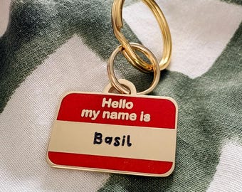 May include: A rectangular gold-toned metal keychain featuring a red and white enamel name tag design. The text "Hello my name is" is embossed in gold, and the name "Basil" is written in black script. It includes a gold-toned split ring attachment.