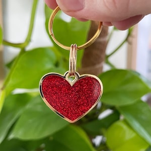 May include: A gold-colored keychain with a heart-shaped charm. The charm has a red glittery surface and a gold-colored border. The keychain is held in front of a background of green leaves.