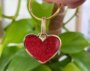 May include: A gold-colored keychain with a heart-shaped charm. The charm has a red glittery surface and a gold-colored border. The keychain is held in front of a background of green leaves.