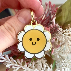 May include: Gold-tone metal keychain featuring a white daisy with a yellow smiling sun face in the centre.
