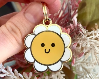 May include: Gold-tone metal keychain featuring a white daisy with a yellow smiling sun face in the center.