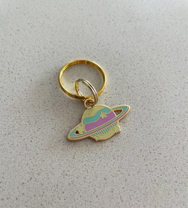May include: A gold-colored keychain with a planet charm. The charm features a light yellow planet with a teal and pink ring, and teal and pink accents. The keychain ring is gold and silver.
