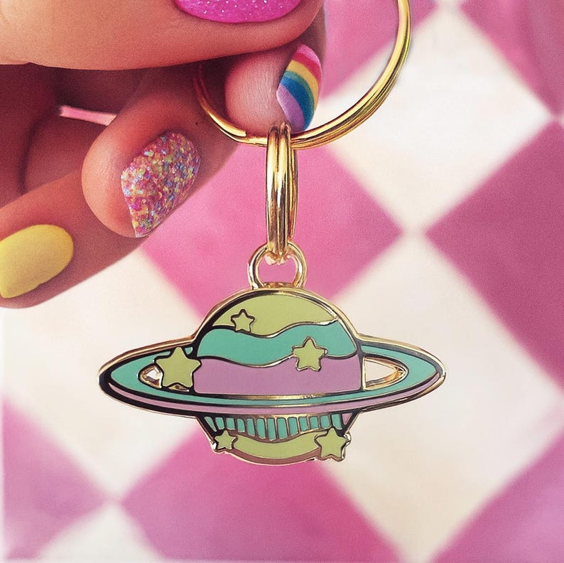 May include: Gold-toned keychain featuring a colorful planet charm. The planet has a light green, pink, and teal design with star accents. The background is a pink and white checkered pattern.