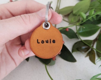 May include: A round, tan leather keychain with a silver ring and the name "Louie" engraved on it.