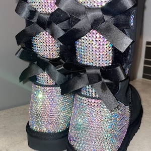 May include: A pair of black boots featuring iridescent rhinestone embellishments covering the back panels. Each boot includes two black satin ribbon bows tied at the rear, black fur lining at the top opening, and a black rubber sole.