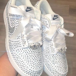 Może przedstawiać: Białe skórzane buty sportowe z białą wstążką satynową i kryształkami, które pokrywają całe obuwie. Logo Nike jest widoczne z boku każdego buta.
