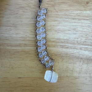 May include: A beaded keychain with a silver-tone chain and clasp. The keychain features a series of clear, round beads connected by a chain, with a rectangular, white stone pendant at the bottom. The background is a light brown wooden surface.