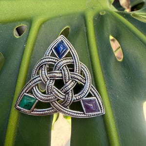 May include: Silver-toned Celtic knot brooch with three colored gemstone accents. The brooch features a central interwoven design with a blue, green, and purple gemstone. The brooch is set against a green leaf.