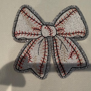 May include: A bow-shaped patch featuring a white fabric base with red stitched baseball seam patterns. The entire edge of the bow is outlined with a thick, shimmering silver glitter border, creating a distinct contrast against the white centre.