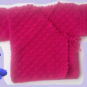 Peut inclure: Un cardigan de bébé au crochet rose vif, fait à la main, avec un design enveloppant. Le cardigan présente un motif de point texturé et une fermeture à nouer. Des décorations de poissons bleus sont éparpillées.