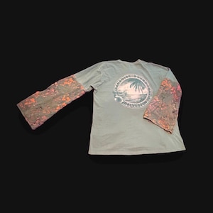 May include: A light green long-sleeve shirt featuring patterned sleeves with orange and dark gray floral accents. The back displays a circular graphic with a palm tree, a beach scene, and the text Paradise Bound Galveston Island.