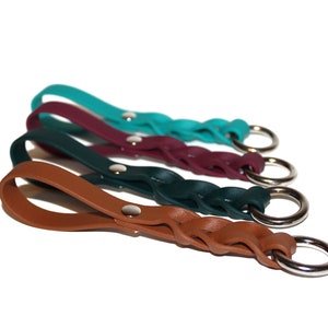 Op de afbeelding: Vier gevlochten leren sleutelhangers in teal, bordeauxrood, donkergroen en bruin met zilveren sleutelringen.