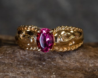 May include: A gold ring with an oval, magenta gemstone. The ring has an ornate, textured design with a braided pattern on the sides. The ring is set on a rough, brown surface.