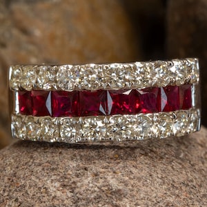 Può includere: Un anello a fascia larga caratterizzato da una fila centrale di cinque rubini rosso intenso a taglio quadrato, affiancati da due file esterne di diamanti bianchi a taglio brillante rotondo. La montatura è realizzata in metallo color argento lucido.