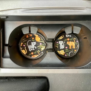 May include: Two black and white cup holders with a cat print design. The cup holders have a black background with white cats, orange cats, and flowers. The text "Meow" is printed on the cup holders.