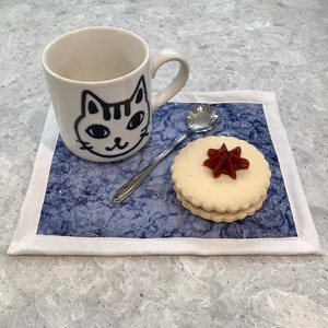 May include: A white ceramic mug with a black cat face design, a silver spoon, and a cookie with red jam on a blue and white patterned fabric coaster.