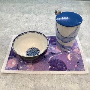 May include: A white ceramic teacup with a blue and white floral design and a blue lid. The teacup is sitting on a purple and white patterned placemat with stars.
