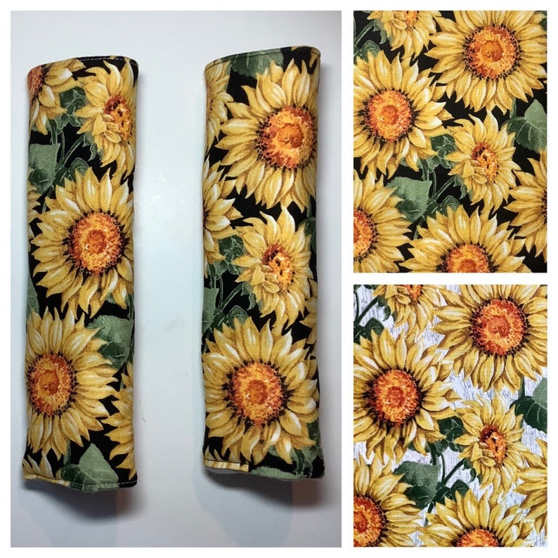 K&ouml;nnte beinhalten: Zwei schwarze Sitzgurtschoner aus Stoff mit Sonnenblumenmuster. Der Stoff hat einen schwarzen Hintergrund mit gelben Sonnenblumen und gr&uuml;nen Bl&auml;ttern.