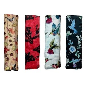 May include: Four fabric seatbelt covers with different patterns. The first cover has a brown and beige background with a bird and leaf design. The second cover has a red background with a black crow design. The third cover has a light blue background with a hummingbird design. The fourth cover has a black background with a hummingbird and floral design.