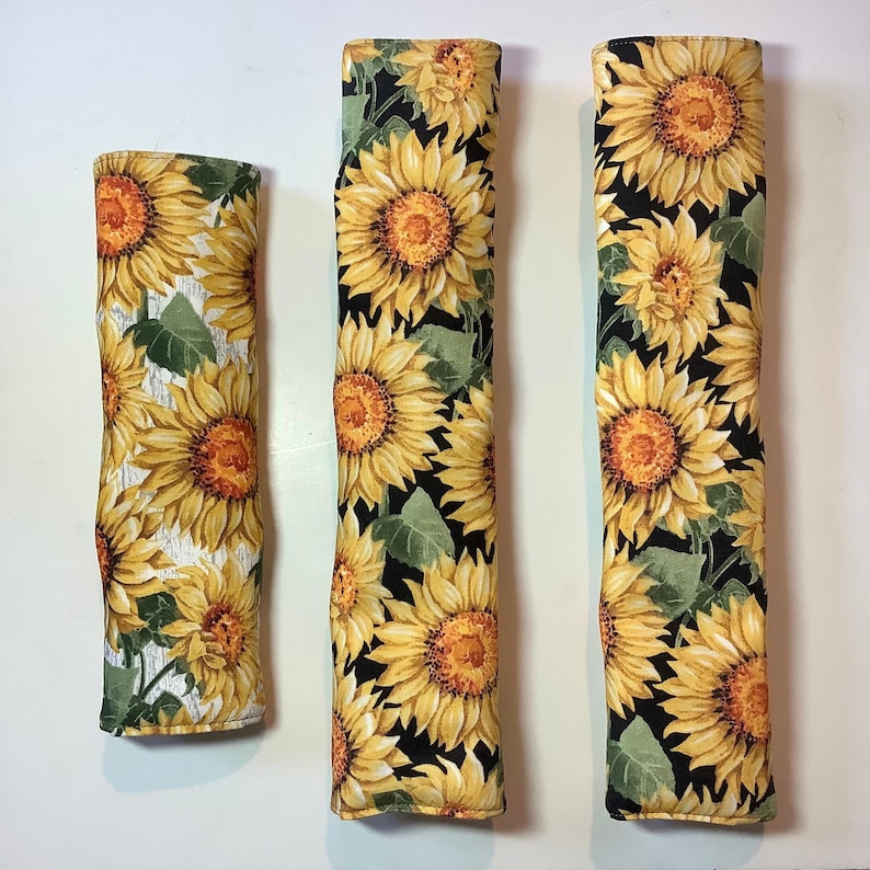 K&ouml;nnte beinhalten: Drei mit Stoff bezogene Ofenhandschuhe mit einem Sonnenblumenmuster. Die Handschuhe sind schwarz mit gelben Sonnenblumen und gr&uuml;nen Bl&auml;ttern. Die Handschuhe sind in verschiedenen Gr&ouml;&szlig;en.