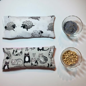 May include: Two gray and white fabric eye pillows. One pillow has a pattern of black and white sheep. The other pillow has a pattern of black and white cats. Both pillows are filled with lavender and flax seeds.