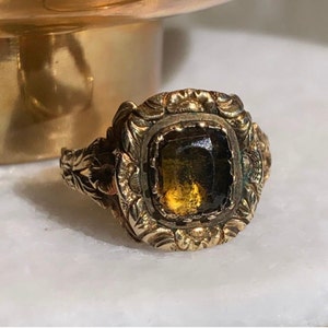 Puede incluir: Anillo dorado ornamentado con una gema cuadrada de color ámbar. El anillo presenta diseños florales y de volutas detallados alrededor de la gema y en la banda. Fondo borroso.