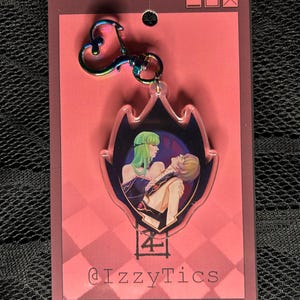 May include: A clear acrylic keychain with a colorful iridescent clasp. The keychain features a stylized image of two figures with green and blonde hair. The background is a dark blue with a pink and red card backing. The text "@IzzyTics" is visible.