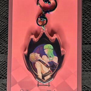 May include: A clear acrylic keychain with a heart-shaped clasp. The keychain features a colorful illustration of two anime characters. The background is a pink card with a checkered pattern and the text "@IzzyTics".