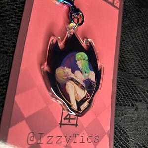 May include: A clear acrylic keychain with a flame-shaped design. The keychain features a colorful illustration of two anime characters. The keychain has a rainbow-colored heart-shaped clasp and the text "@IzzyTics".