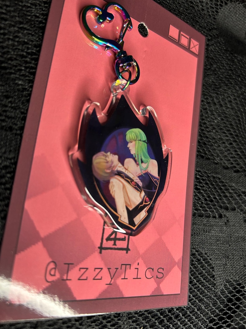 May include: A clear acrylic keychain with a flame-shaped design. The keychain features a colorful illustration of two anime characters. The keychain has a rainbow-colored heart-shaped clasp and the text "@IzzyTics".